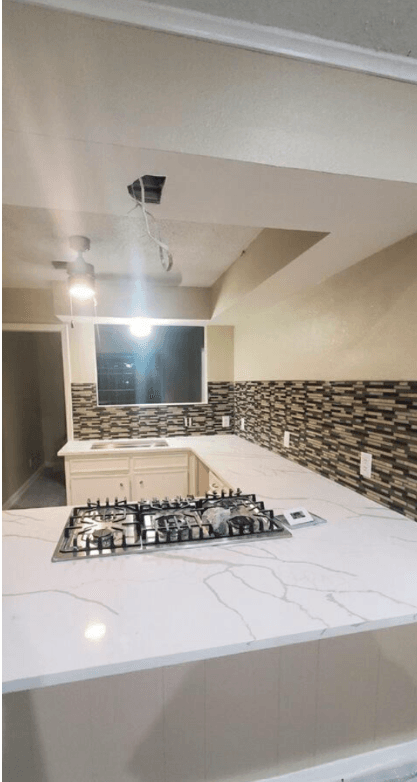 Kitchen peninsula topped in white marble-look quartz with a stainless steel gas cooktop set into it, against a mosaic tile backsplash.
