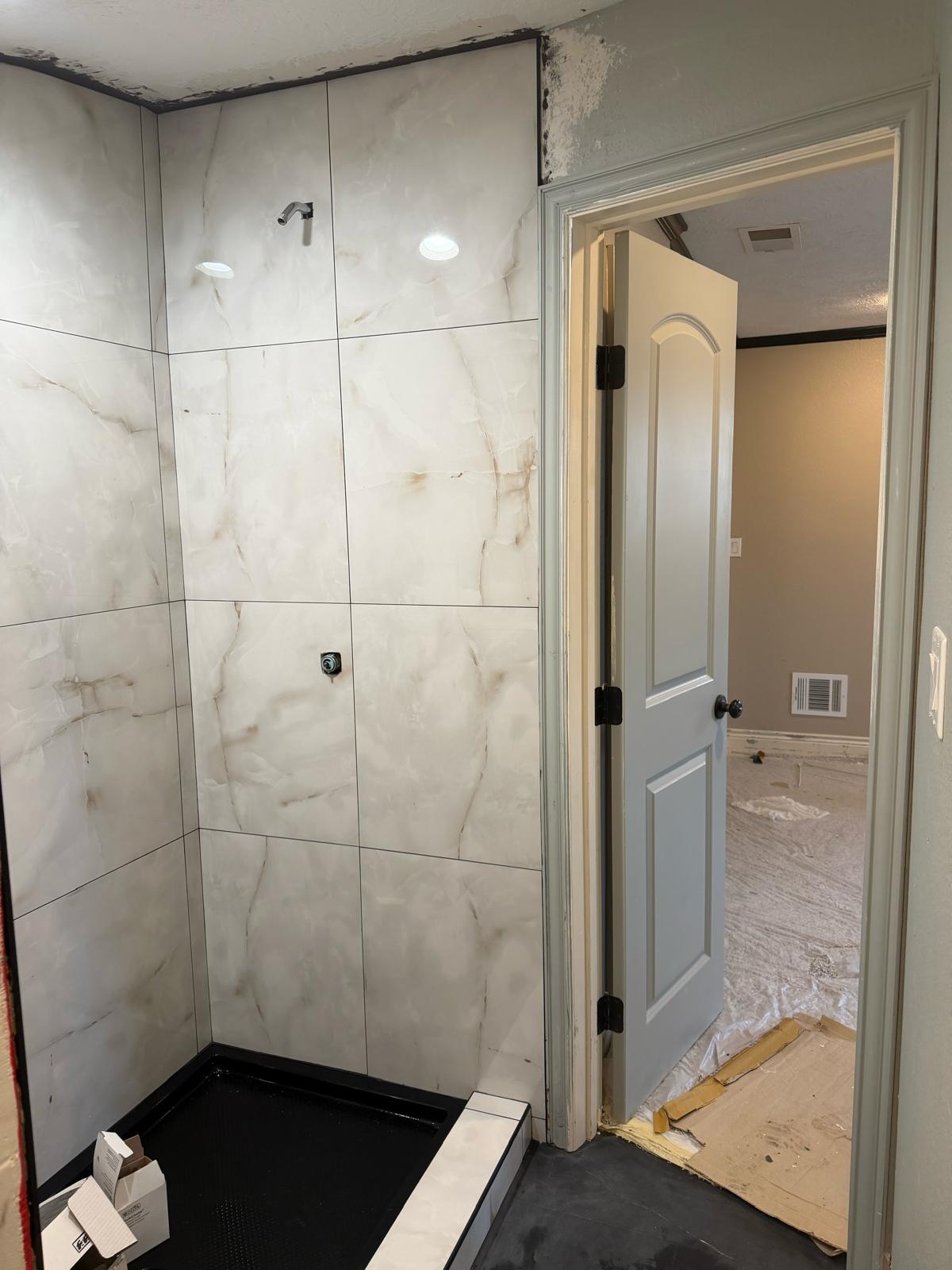 Walk-in shower lined with large-format marble-look porcelain tile, with a black shower pan and recessed lights, in a bathroom still being remodeled.