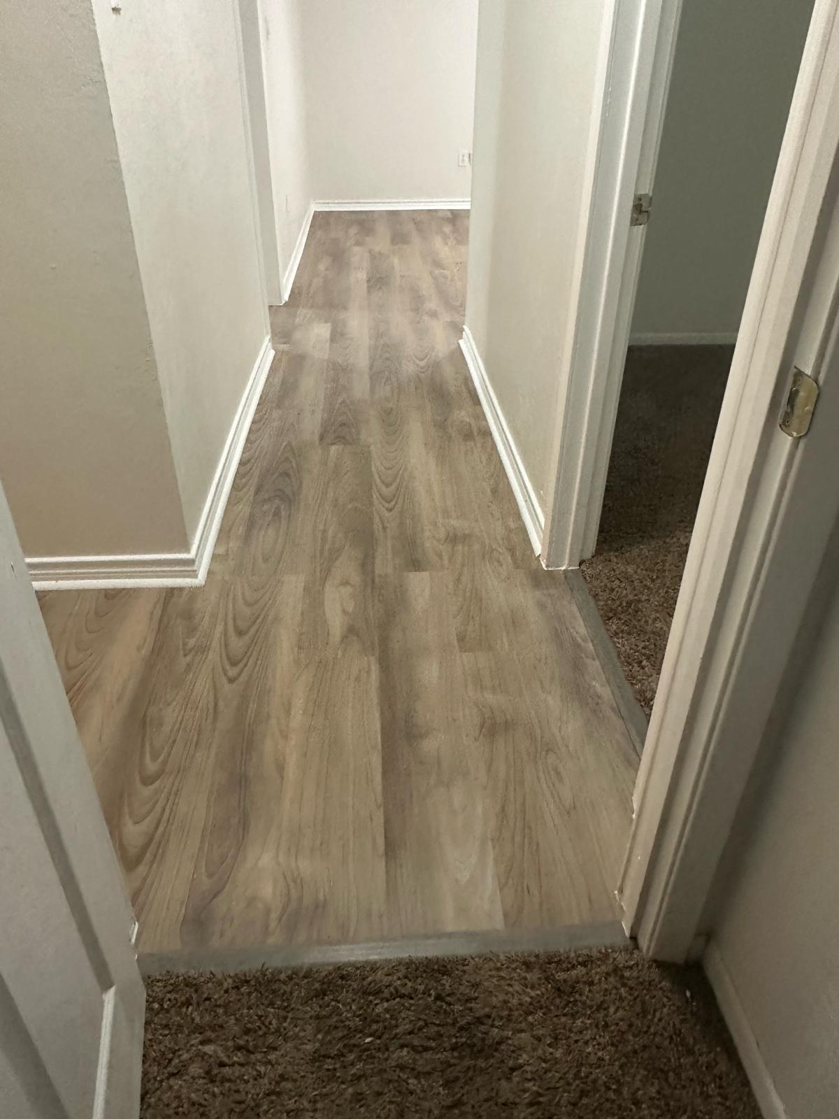Warm gray-brown wood-look plank flooring newly laid down a hallway, with white baseboards and door frames, meeting brown carpet at the doorway thresholds.