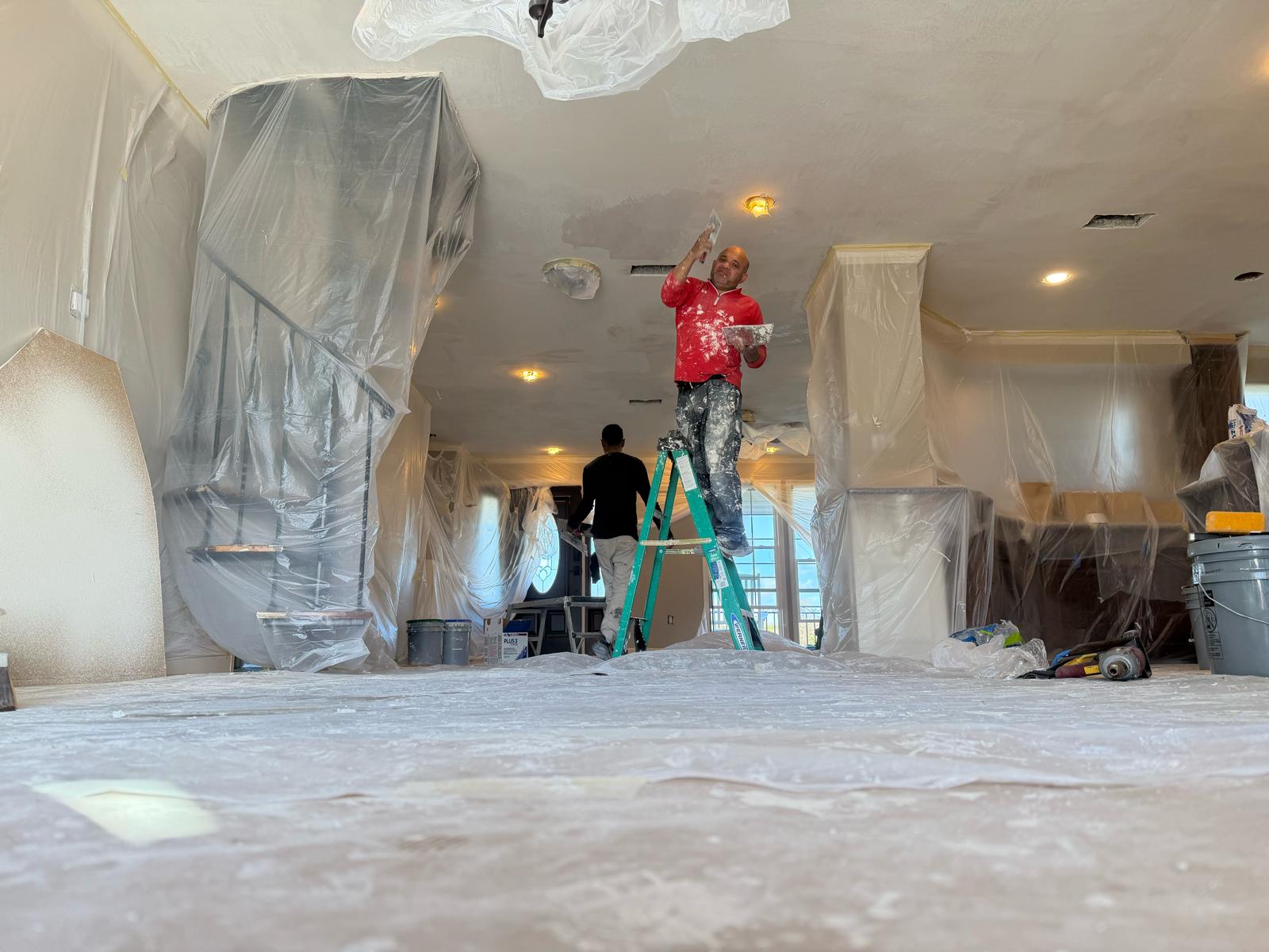 A crew member on a step ladder smoothing joint compound onto a ceiling with a taping knife, in a room where the stair rail, furniture and floor are all covered in plastic sheeting.
