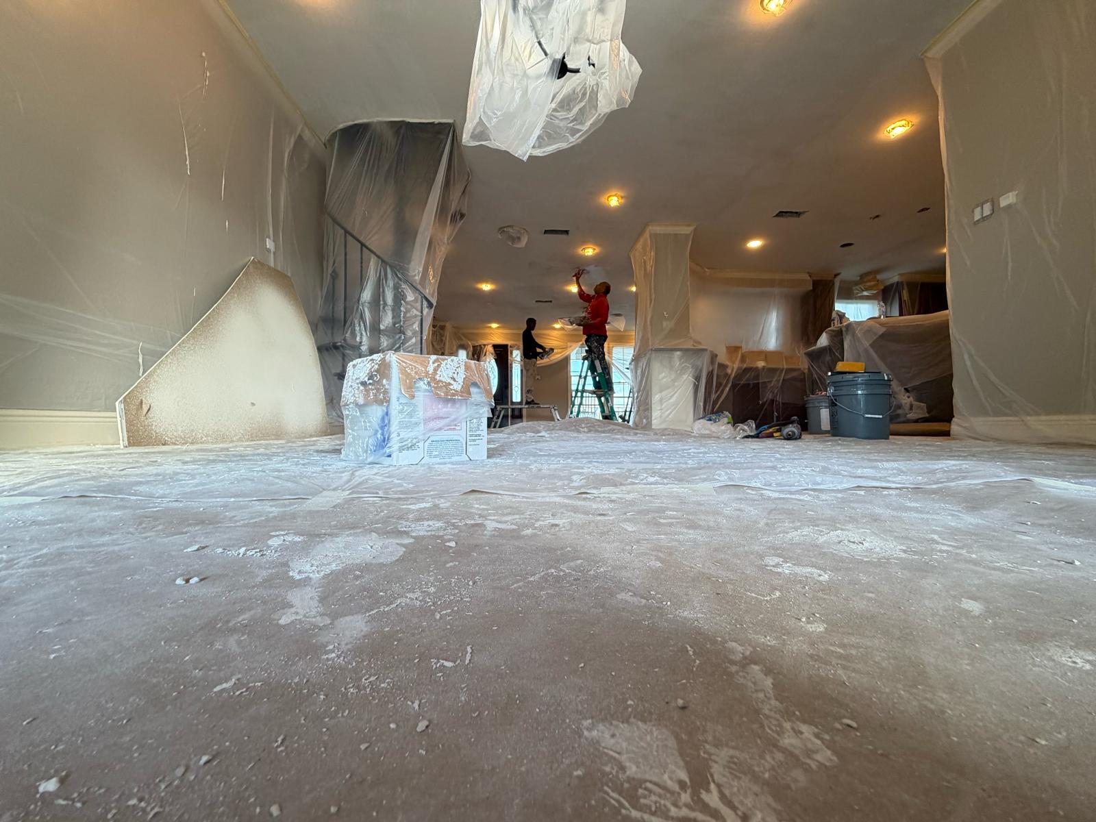 Wide view of a living area fully masked in plastic sheeting — floor, staircase and furniture all covered — while two crew members work on the ceiling at the far end.